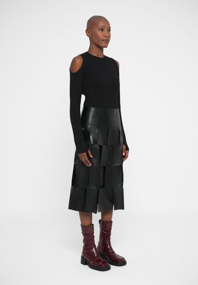 Mujer con una blusa negra de hombros al descubierto, una falda de cuero negra en capas con paneles rectangulares y botas brillantes de color rojo oscuro hasta la mitad de la pantorrilla, de pie de perfil.