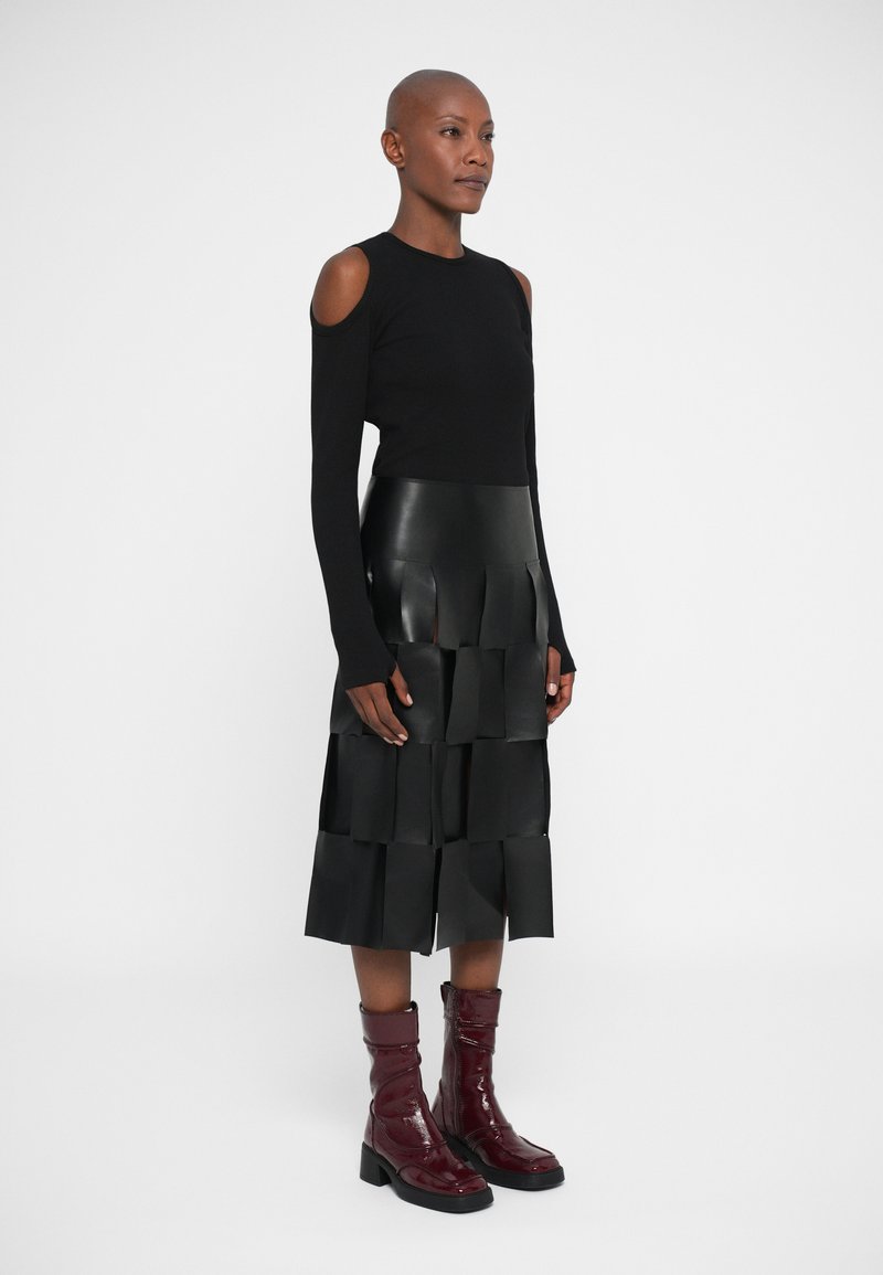 Vrouw gekleed in een zwart cold-shoulder top, een gelaagde zwarte leren rok met rechthoekige panelen en glanzende donkerrode laarzen tot halverwege het kuit, staande in zijaanzicht.