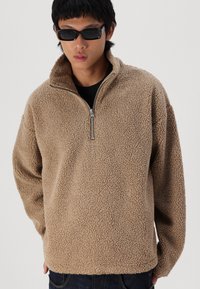 Bruine fleece trui met een hoge kraag en ritssluiting. Heeft een gestructureerd oppervlak en een losse fit, gedragen over een zwart shirt.