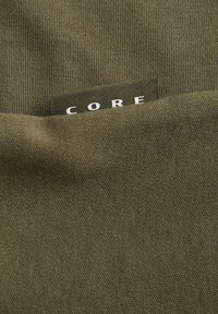 Tela verde oliva con una superficie texturizada, que presenta una etiqueta negra con la palabra "CORE" en letras blancas.