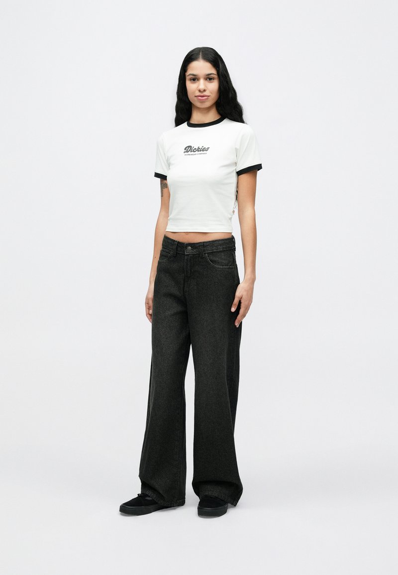 Junge Frau mit langen dunklen Haaren, die ein weißes, bauchfreies T-Shirt mit schwarzem Besatz und schwarze Jeans mit weitem Bein trägt und vor weißem Hintergrund steht.