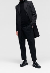 Černý kabát s klopami, zapínání na knoflíky, nošený přes černý rolák. Černé kalhoty a robustní černé boty dotvářejí celkový vzhled.