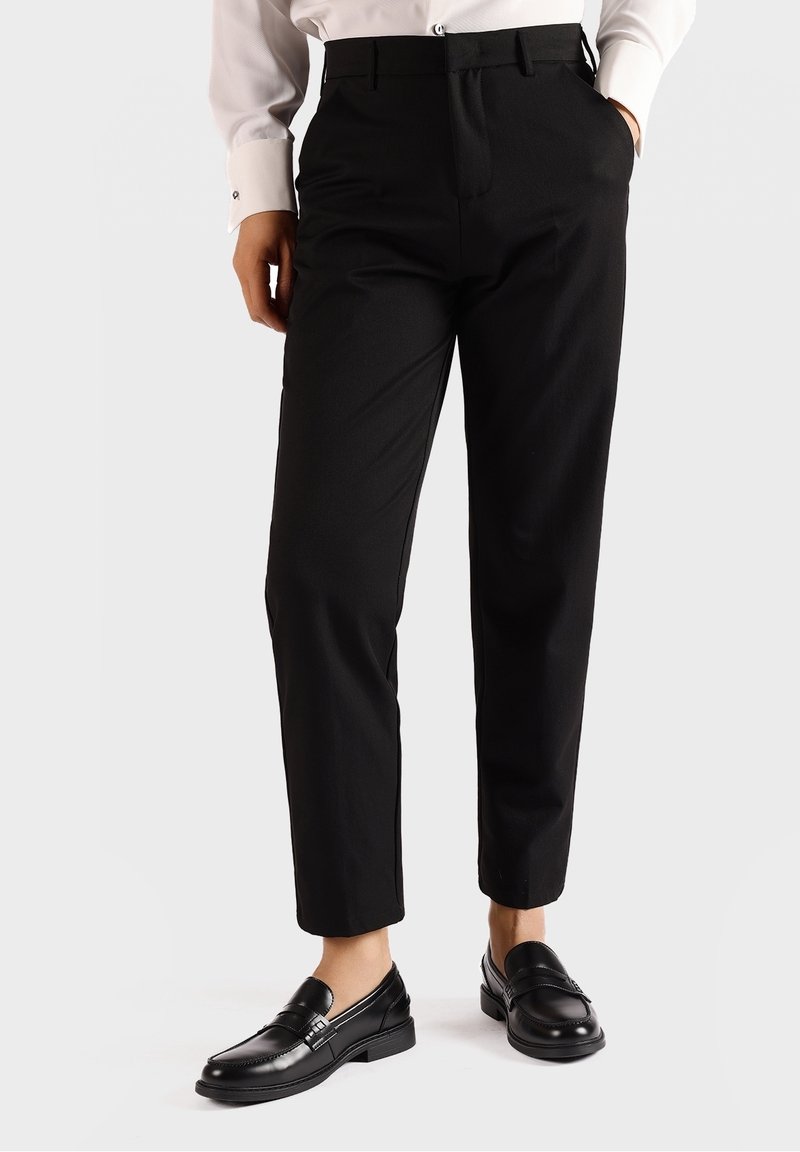 Personne portant un pantalon noir sur mesure avec des poches à l'avant et des mocassins en cuir noir, associé à une chemise blanche à manches longues.