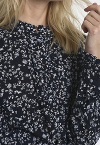 Granatowa bluzka z wzorem w kwiaty w białym i jasnoniebieskim kolorze, z wysokim falbaniastym dekoltem i guzikami z przodu, wykonana z miękkiego materiału.