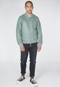 Bledě zelená kožená bomberka s žebrovanými manžetami a lemem, přední zipové kapsy, nošena s tmavými roztrhanými džíny a hnědými vysokými teniskami.