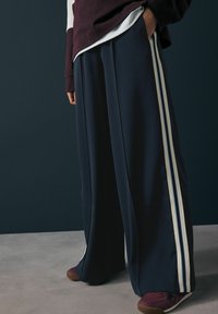 Pantalons larges bleu marine avec des rayures blanches et bleu clair sur les côtés, fabriqués à partir d'un matériau doux. Associés à des baskets de couleur bordeaux foncé.