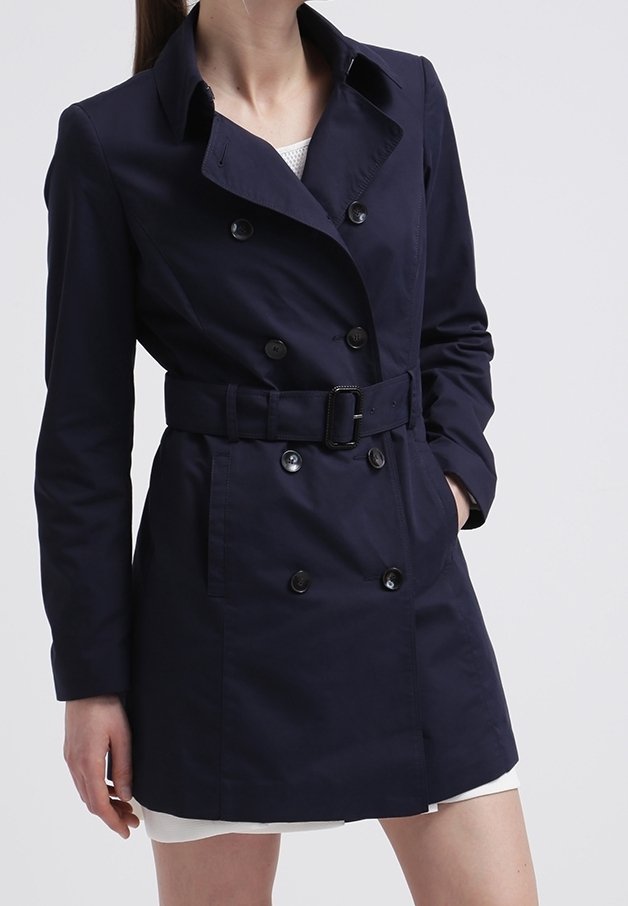 Vrouw draagt een marineblauwe dubbelgeknoopte trenchcoat met riem over een witte jurk, hand in de zak, staande tegen een effen achtergrond.