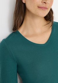 Teal pitkähihainen paita, jossa on V-pääntie. Kangas vaikuttaa pehmeältä ja sileältä, eikä siinä ole näkyviä kuvioita tai koristeluita.
