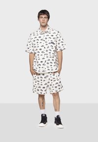 Set di camicia bianca a maniche corte e pantaloni corti con un motivo di occhi neri. Il materiale sembra leggero. Indossato con sneakers nere e calzini bianchi.