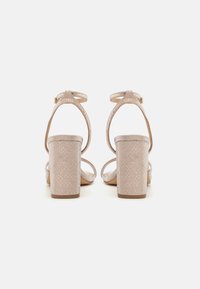 Une paire de sandales à talons blocs ouvertes en beige avec des brides de cheville et une texture de serpent subtile, présentée de dos sur un fond blanc.