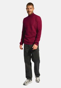 Burgundský rolák vyrobený z texturované tkaniny, v kombinaci s černými džíny a teniskami s bílým a šedým designem. Model stojí v uvolněné pozici.