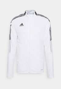 Hvid Adidas trackjakke med sorte tre-stribe accenter på ærmerne, lynlås foran, høj krave og glat stoftekstur.