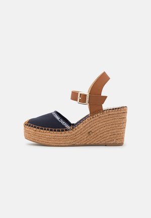 Navy canvas wedge sandal with brown adjustable strap, woven jute heel, and contrasting stitching detail. Closed toe design with a decorative logo.