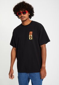 Black cotton t-shirt featuring a small graphic in orange, yellow, and black on the left side; paired with red sunglasses and blue jeans.