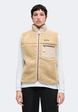 Persona con capelli corti e scuri indossa un gilet in pile beige con tasca zip sul petto sopra una camicia bianca a maniche lunghe e pantaloni neri, rivolta in avanti.
