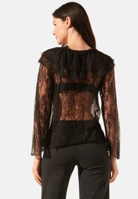 Blouse en dentelle noire avec des manches longues, un col en volants et un dos transparent, ornée de motifs floraux complexes et d'une coupe ajustée.