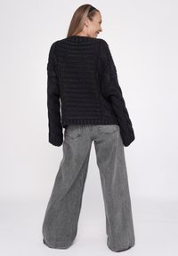 Schwarzer, grob gestrickter Pullover mit weiten Ärmeln und rundem Ausschnitt, kombiniert mit lockeren, grauen, hochtaillierten Jeans. Schlichter weißer Hintergrund.