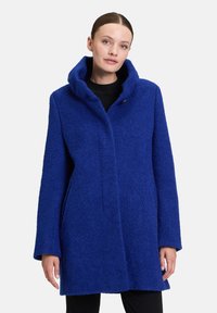 Manteau en mélange de laine bleu avec un col montant, poches latérales et une fermeture à bouton unique. Tissu texturé et chaud, longueur sous la hanche.