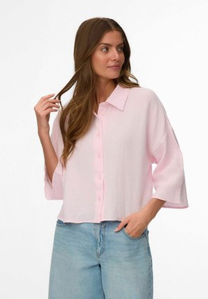 Kobieta o długich brązowych włosach, ubrana w luźną, jasnoróżową koszulę na guziki z rękawami 3/4 oraz jasnoniebieskie dżinsy, stojąca z jedną ręką w kieszeni.