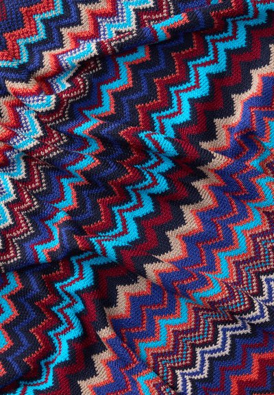 Manta de malha com um padrão em ziguezague nas cores vermelho, azul, bege e azul-marinho. Textura suave com um entrelaçamento denso, destacando um vibrante contraste de cores.
