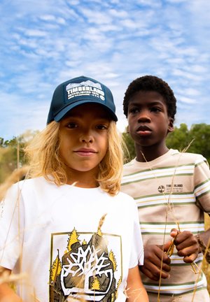 Due bambini stanno in un campo. Uno indossa un berretto blu scuro e una maglietta bianca con una grafica, l'altro una maglietta beige a righe, con un ciuffo d'erba in mano.