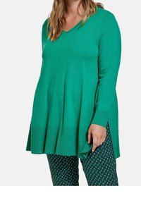 Zelený svetr s dlouhým rukávem a véčkovým výstřihem, strukturovaným žebrovaným lemem a postranními rozparky, v kombinaci s vzorovanými zelenými kalhotami s diamantovými tvary.