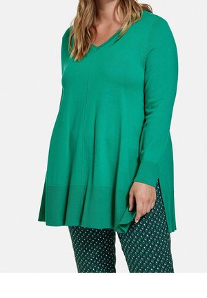 Vihreä pitkähihainen neulepaita V-kauluksella, teksturoidulla resorilla helmassa ja sivuhaaroilla, yhdistetty kuvioituihin vihreisiin housuihin, joissa on timantinmuotoisia kuvioita.