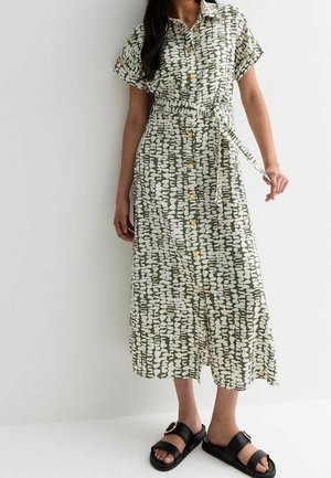 Femme portant une robe-chemise midi à motifs verts et blancs avec une ceinture en tissu et des sandales noires à boucle, debout contre un mur blanc.