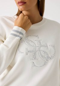 Maglione bianco a maglia con un grande logo testurizzato e dettagli brillanti. Le maniche a coste a righe grigie completano il design.