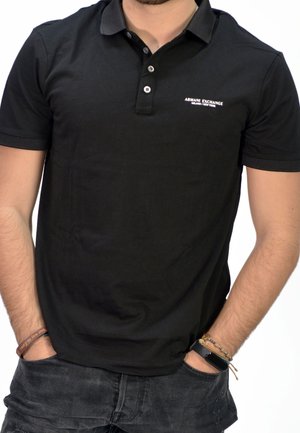 Black polo shirt made of soft fabric with a classic collar, three buttons, and a small white logo on the chest. Worn with dark jeans.