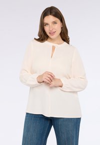 Blusa color pesca chiaro con scollo rotondo, dettaglio a chiave e maniche lunghe elasticizzate, abbinata a pantaloni in denim a righe blu.