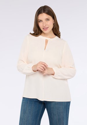 Helles Pfirsichbluse mit runder Halslinie, Schlüsselverschluss-Detail und elastischen langen Ärmeln, kombiniert mit blauen gestreiften Denim-Hosen.