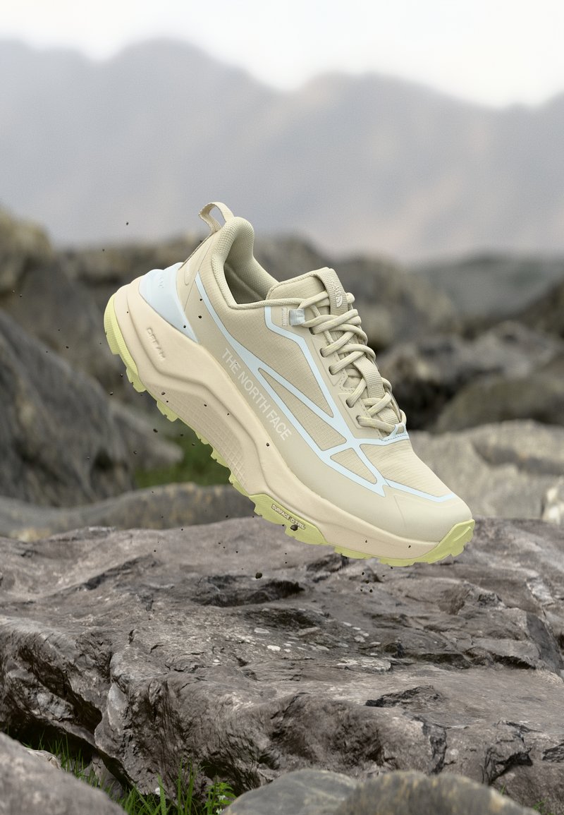 Ανοιχτό μπεζ πεζοπορικό παπούτσι The North Face κρεμασμένο πάνω από βραχώδες έδαφος με θολό φόντο βουνών.