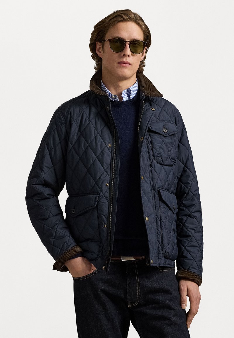 Quilted marineblauer Jacke mit braunem Cordkragen, Vordertaschen und einem Reißverschluss, getragen über einem marineblauen Pullover und einem blau gestreiften Hemd.