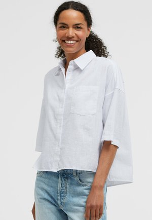 Lächelnde Frau mit lockigem Haar, die ein lockeres weißes Hemd mit Knöpfen und hellblaue Jeans vor einem schlichten Hintergrund trägt.