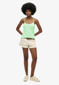 Top verde sin mangas con tirantes finos, combinado con shorts desgastados beige claros y zapatillas ligeras, todo sobre un fondo blanco.