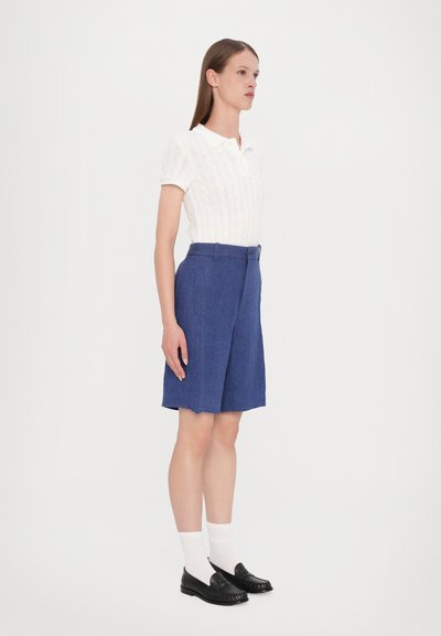 Polo shirt blanc à manches courtes et texturé ; short bleu ajusté avec une fermeture à bouton ; porté avec des mocassins noirs et des chaussettes blanches.