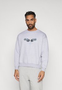 Sweatshirt cinza claro com gola redonda, ombros caídos e punhos canelados, apresentando uma estampa azul e preta no peito.