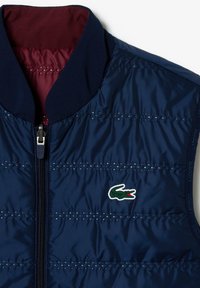 Chaleco acolchado azul marino con cremallera frontal, cuello oscuro, forro interior burdeos y pequeño logo de cocodrilo verde en el pecho.