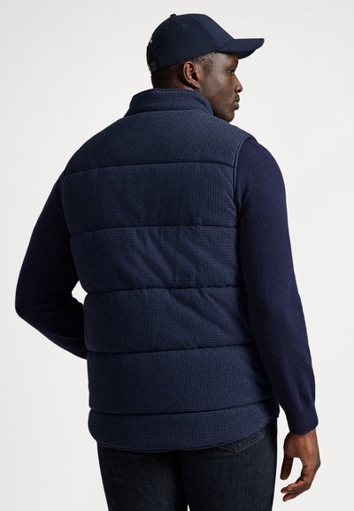 Granatowa kamizelka puchowa z teksturowanej tkaniny, z pionowym pikowaniem, wysokim kołnierzem i kontrastowymi dzianinowymi rękawami. Noszona z dopasowaną czapką.
