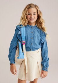 Blaue Denim-Bluse mit rüschigen Schultern und Knopfverschluss, kombiniert mit einem beigen Faltenrock. Halten einer hellblauen Tasche mit pinken Akzenten.