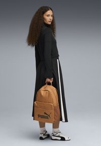Mochila marrón claro con un gran bolsillo frontal y el logo negro de Puma, sostenida por un modelo con un top y una falda negra con rayas blancas.