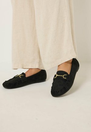 Pieds chaussés de mocassins en daim noir avec détail mors doré, associés à un pantalon ample en lin beige.