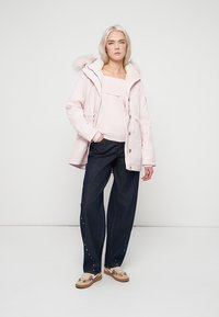 Casaco com capuz rosa e acabamento em pelo, suéter leve e jeans de perna larga azul escuro com detalhes em pérolas, combinados com sapatos estampados.