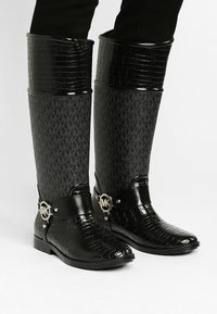Bottes montantes imperméables noires en caoutchouc, présentant un dessus en faux crocodile texturé, un tige en tissu avec un motif de logo, et un détail de boucle métallique.