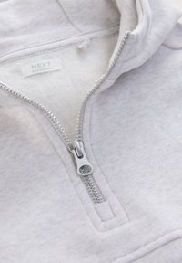 Hellgrauer Kapuzenpullover mit Reißverschluss, glatter Textur, YKK-Reißverschluss und einem kleinen Etikett innen. Verfügt über eine Kapuze und eine Öffnung für die Fronttasche.