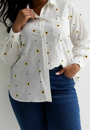 Weißes Hemd mit gelben Sonnenblumenmustern, getragen zu blauer Jeans, eine Hand berührt den Kragen, die andere steckt in der Tasche.