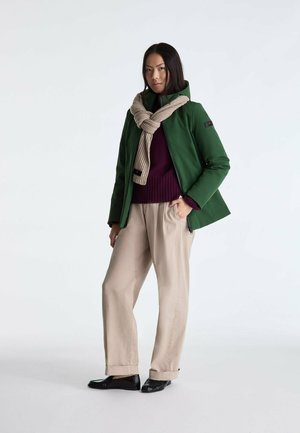 Femme portant une veste verte, une écharpe beige nouée autour du cou, un pull violet, un pantalon beige et des chaussures noires, debout sur un fond blanc.