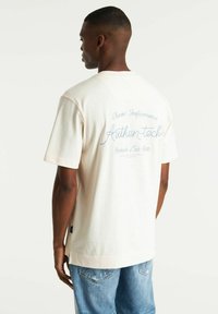 T-shirt a maniche corte color crema con testo ricamato blu sul retro, vestibilità rilassata, in cotone, abbinato a pantaloni corti di jeans blu chiaro.