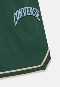 Pantalones cortos verdes hechos de una tela suave, con un logotipo curvado de "CONVERSE" en azul claro. Los bordes tienen acentos en beige y un dobladillo a rayas.